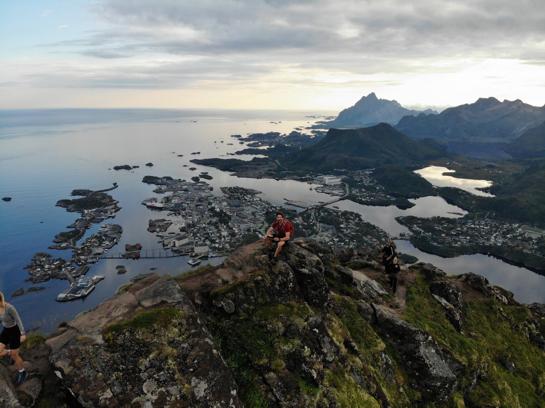 A hiker in a red shirt resting on a summit above Svolvaer in the Lofoten Islands, Norway, with the fjord, harbour town, and jagged peaks below at dusk